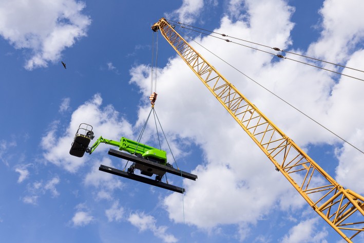 DeckRider being lifted by crane Niftylift DeckRider suspended from a crawler crane against a blue sky, its steel skids visible beneath the machine